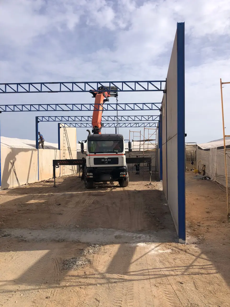 Camión grúa izando cerchas de celosía azules para una nave metálica entre invernaderos