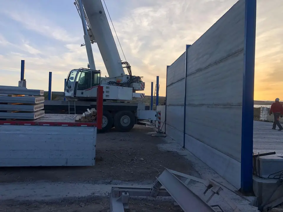 Grúa colocando paneles prefabricados de hormigón entre pilares metálicos azules al atardecer