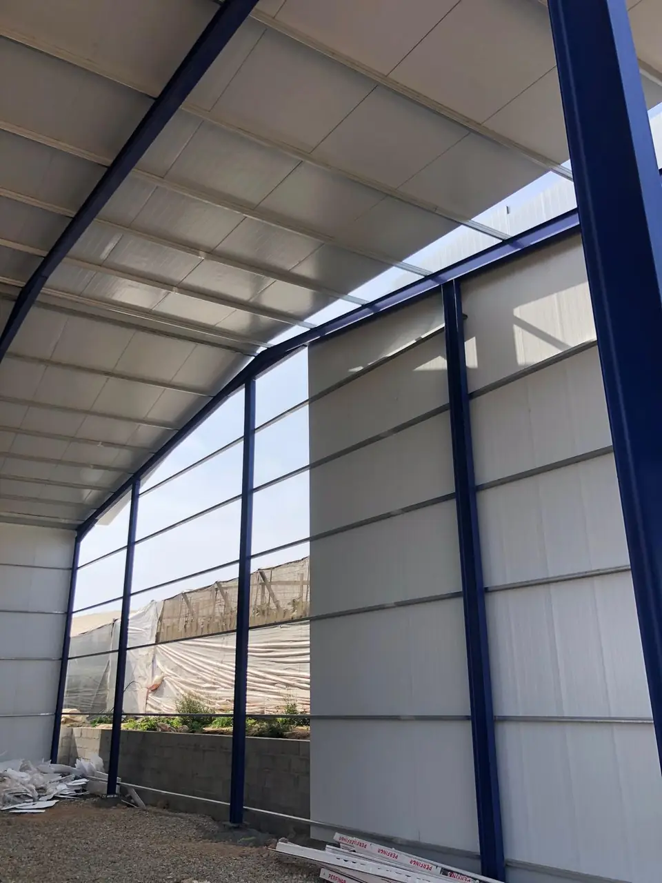 Lateral de nave con pórticos azules y panel sándwich, con invernaderos al fondo